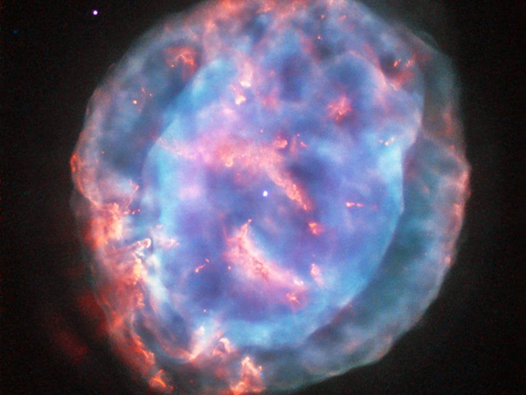This colourful bubble is a planetary nebula called NGC 6818, also known as the Little Gem Nebula. It is located in the constellation of Sagittarius (The Archer), roughly 6000 light-years away from us. The rich glow of the cloud is just over half a light-year across — humongous compared to its tiny central star — but still a little gem on a cosmic scale. When stars like the Sun enter retirement, they shed their outer layers into space to create glowing clouds of gas called planetary nebulae. This ejection of mass is uneven, and planetary nebulae can have very complex shapes. NGC 6818 shows knotty filament-like structures and distinct layers of material, with a bright and enclosed central bubble surrounded by a larger, more diffuse cloud. Scientists believe that the stellar wind from the central star propels the outflowing material, sculpting the elongated shape of NGC 6818. As this fast wind smashes through the slower-moving cloud it creates particularly bright blowouts at the bubble’s outer layers. Hubble previously imaged this nebula back in 1997 with its Wide Field Planetary Camera 2, using a mix of filters that highlighted emission from ionised oxygen and hydrogen (opo9811h). This image, while from the same camera, uses different filters to reveal a different view of the nebula. A version of the image was submitted to the Hubble’s Hidden Treasures image processing competition by contestant Judy Schmidt.